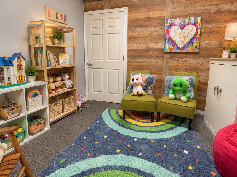Children's play therapy room with a dollhouse, toy shelves, green chairs, and a rainbow rug